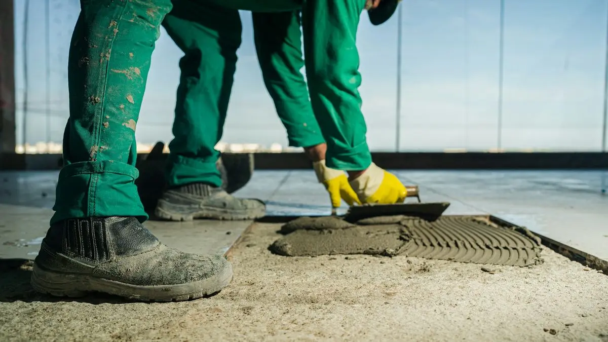 Fliesenleger in grüner Arbeitshose zieht mit der Kelle Kleber auf, die ersten Platten liegen schon – die Arbeit ist halb fertig