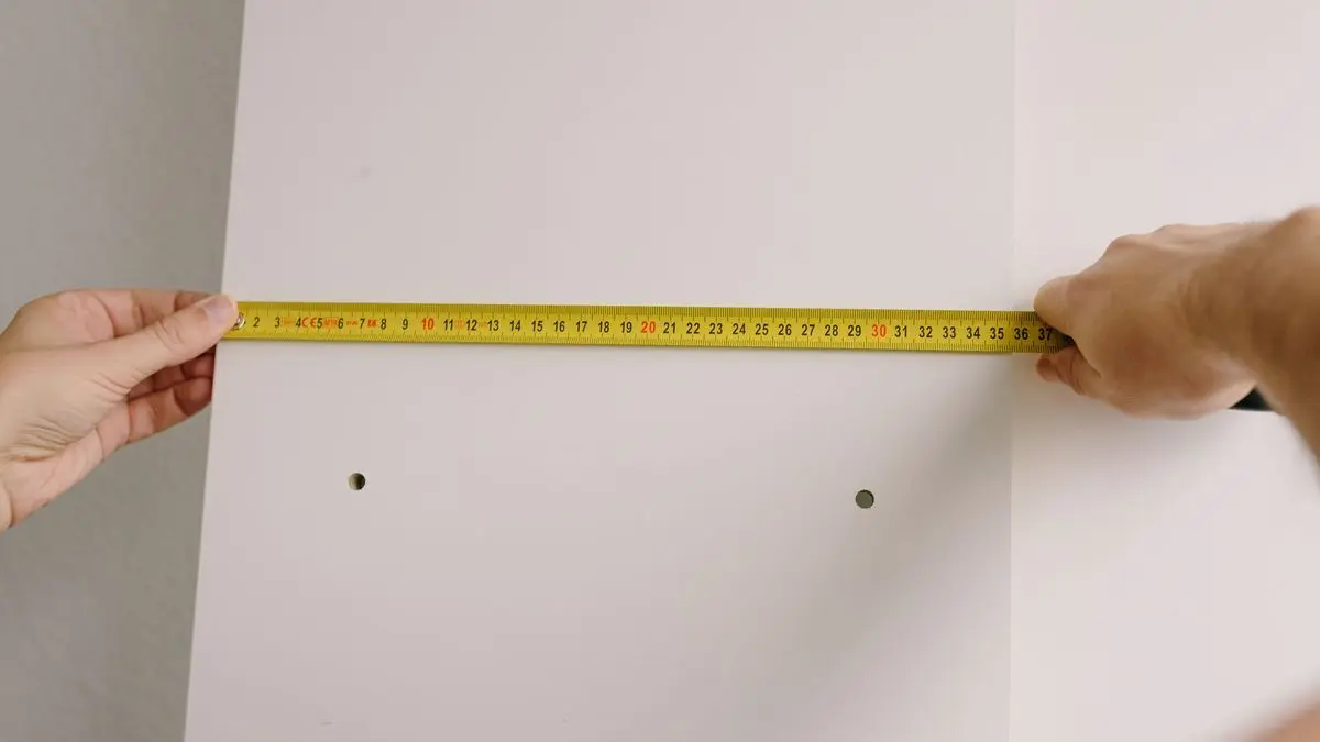 Zwei Hände halten ein gelbes Maßband quer über eine helle Wandplatte