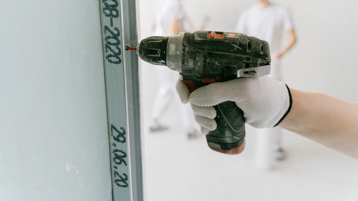 Trockenbauer schraubt mit dem Akkuschrauber eine Gipskartonplatte an ein Metallprofil, im Hintergrund arbeiten weitere Handwerker