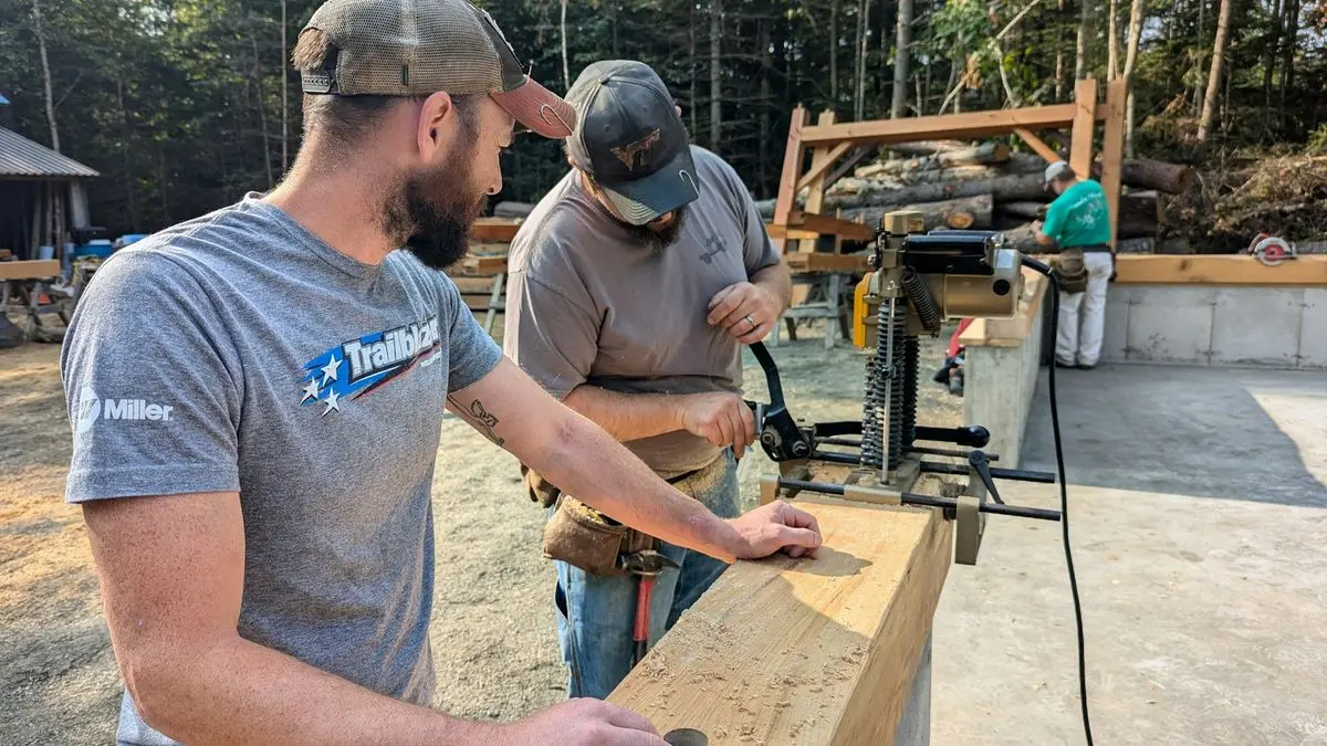 Zwei Handwerker arbeiten draußen gemeinsam an einem Holzbalken, im Hintergrund die Kappsäge