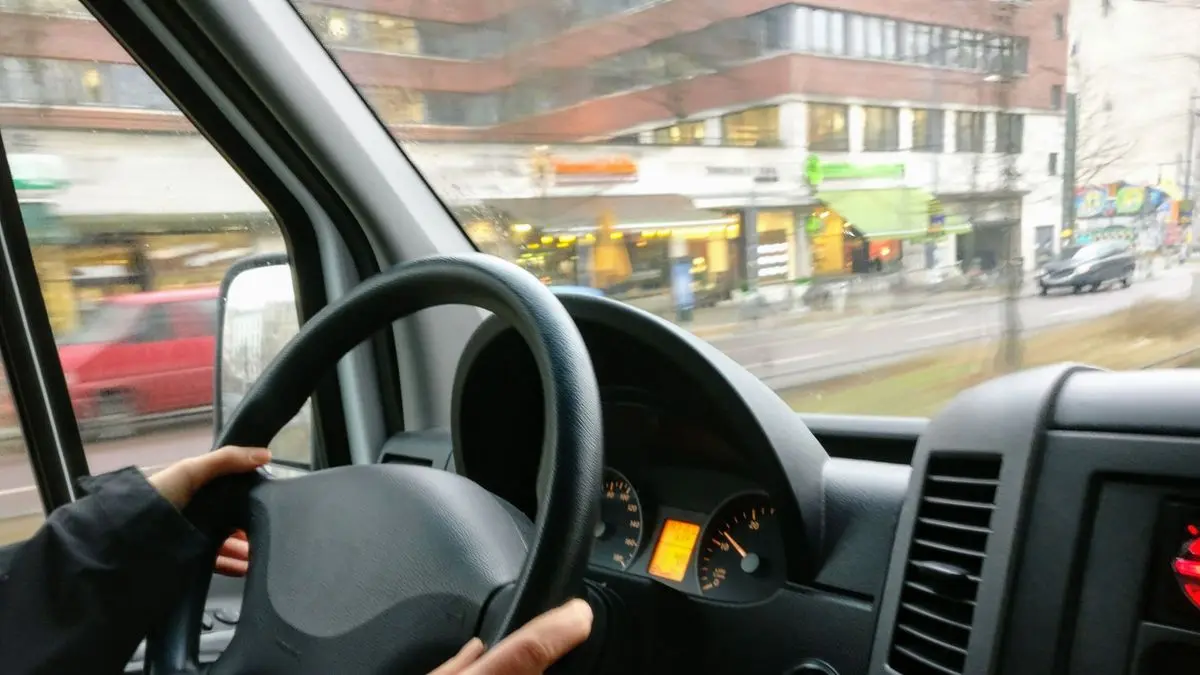 Blick vom Fahrersitz eines Transporters: zwei Hände am Lenkrad, draußen zieht eine Stadtstraße mit Geschäften vorbei