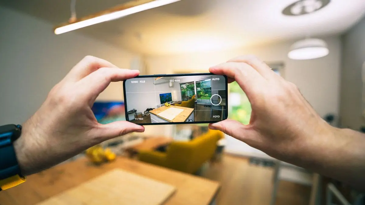 Zwei Hände halten ein Smartphone quer und fotografieren einen Wohnraum