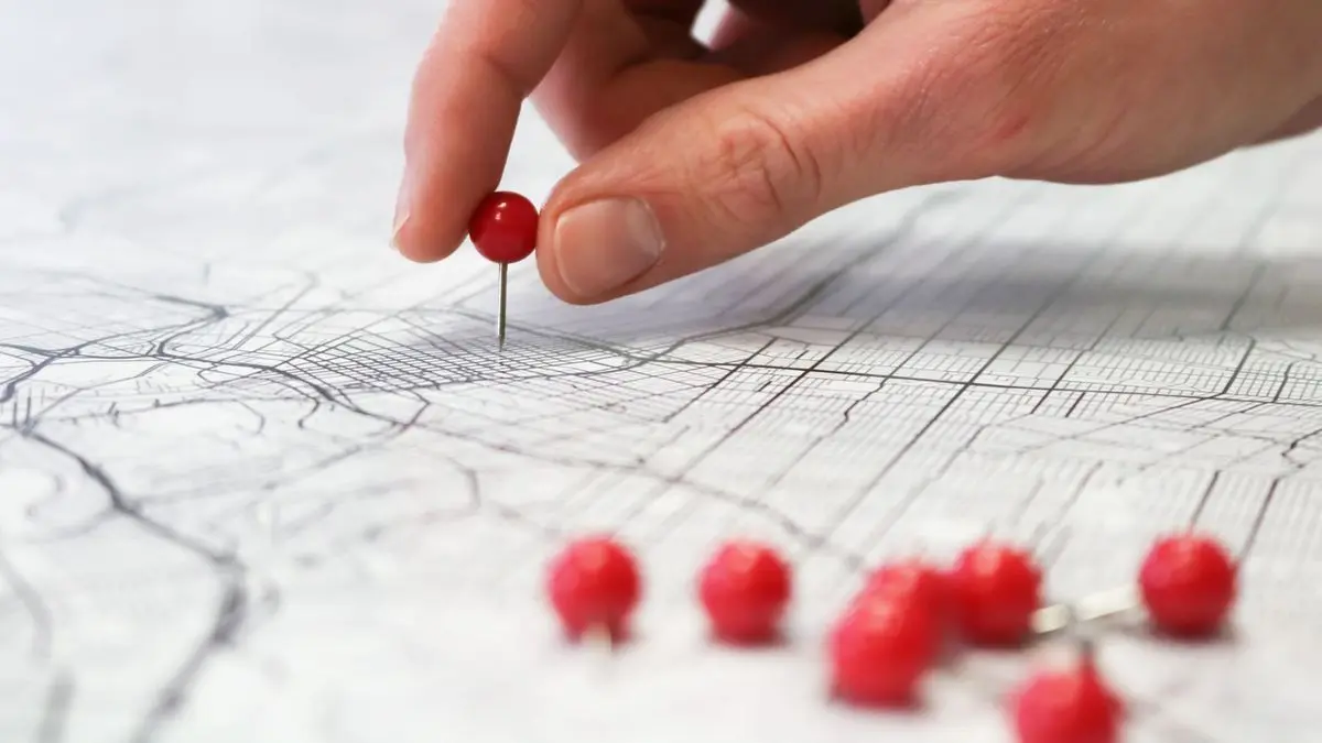 Eine Hand steckt eine rote Stecknadel in einen gedruckten Stadtplan, im Vordergrund liegen weitere rote Nadeln
