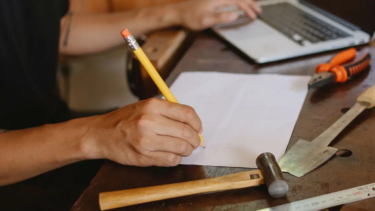 Hand mit Bleistift schreibt an der Werkbank Zahlen auf ein Blatt, daneben liegen Hammer, Säge, Zange und ein Laptop