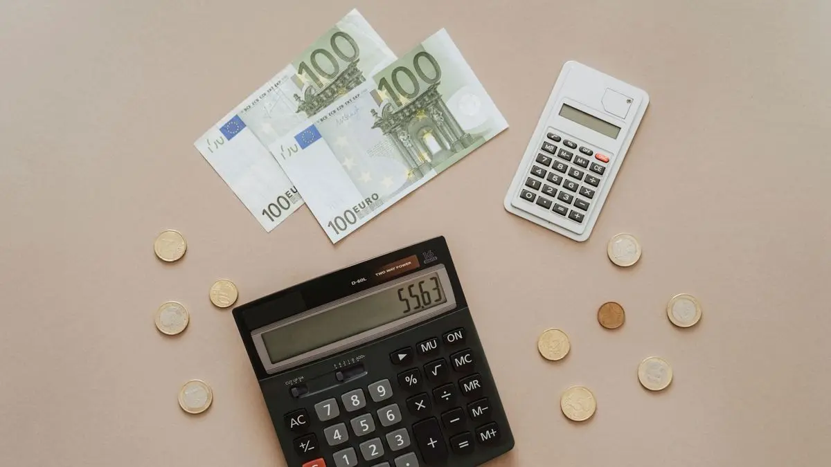 Zwei 100-Euro-Scheine, Euro-Münzen und zwei Taschenrechner auf einem hellbraunen Tisch