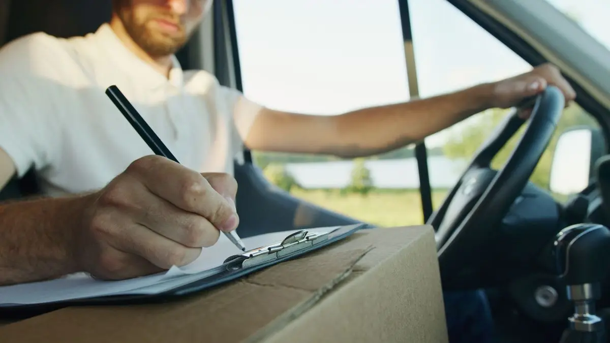 Mann im Transporter schreibt mit einem Stift auf ein Klemmbrett, das auf einem Karton liegt, die andere Hand am Lenkrad