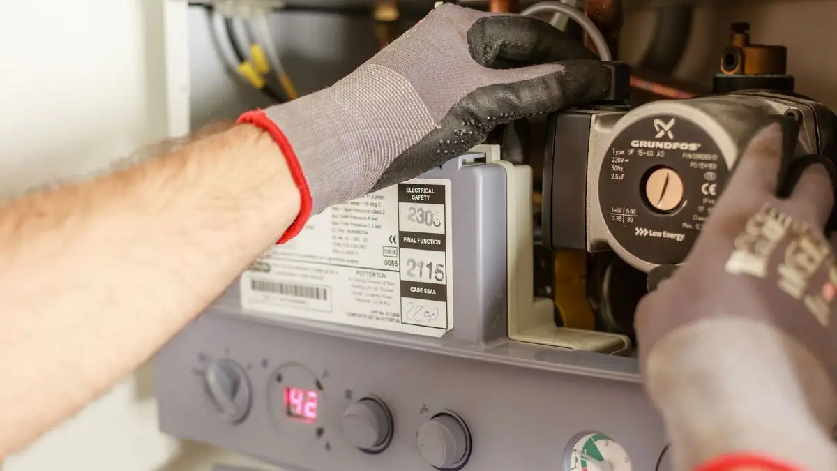 Hände in Arbeitshandschuhen an der geöffneten Therme einer Heizung, daneben Umwälzpumpe, Typenschild und Manometer
