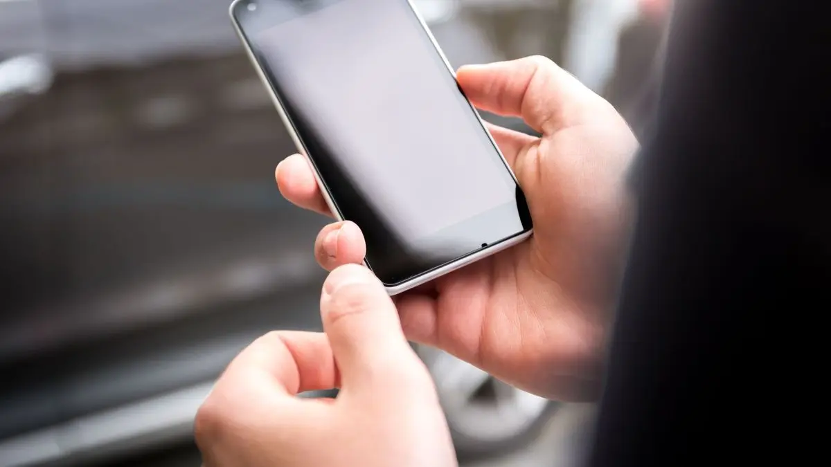 Zwei Hände halten ein Smartphone mit leerem Bildschirm, im Hintergrund unscharf ein Auto