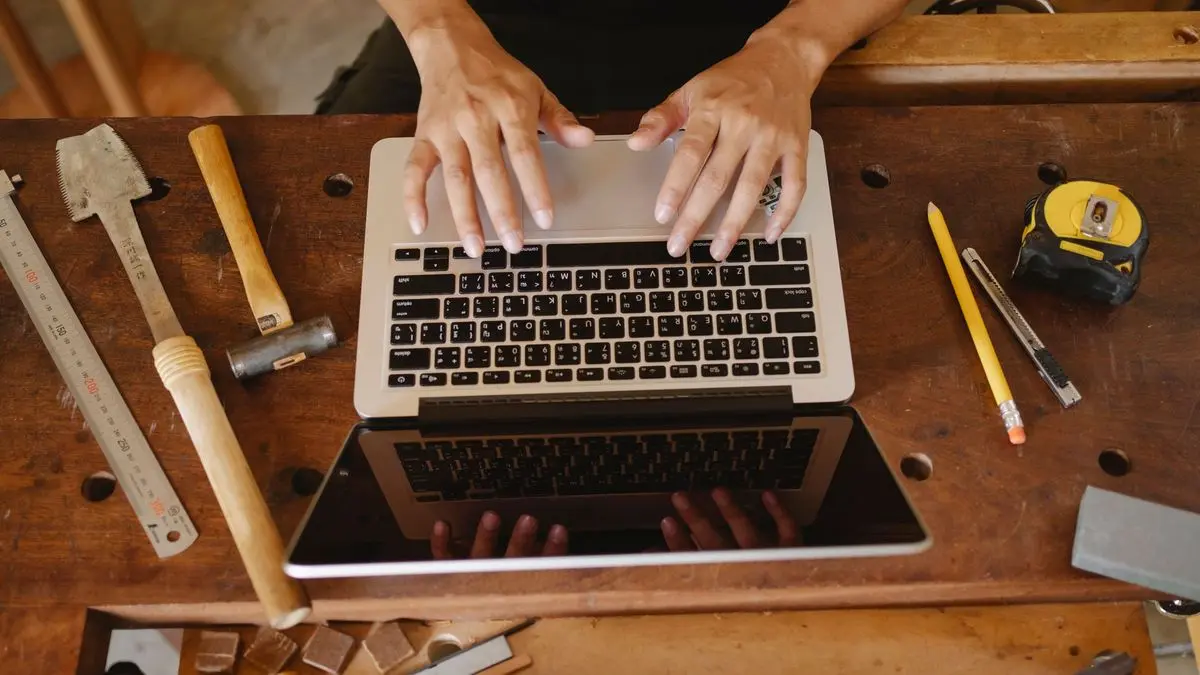 Blick von oben auf eine Hobelbank: Hände tippen auf einem Laptop, daneben liegen Hammer, Maßstab, Bleistift und Maßband