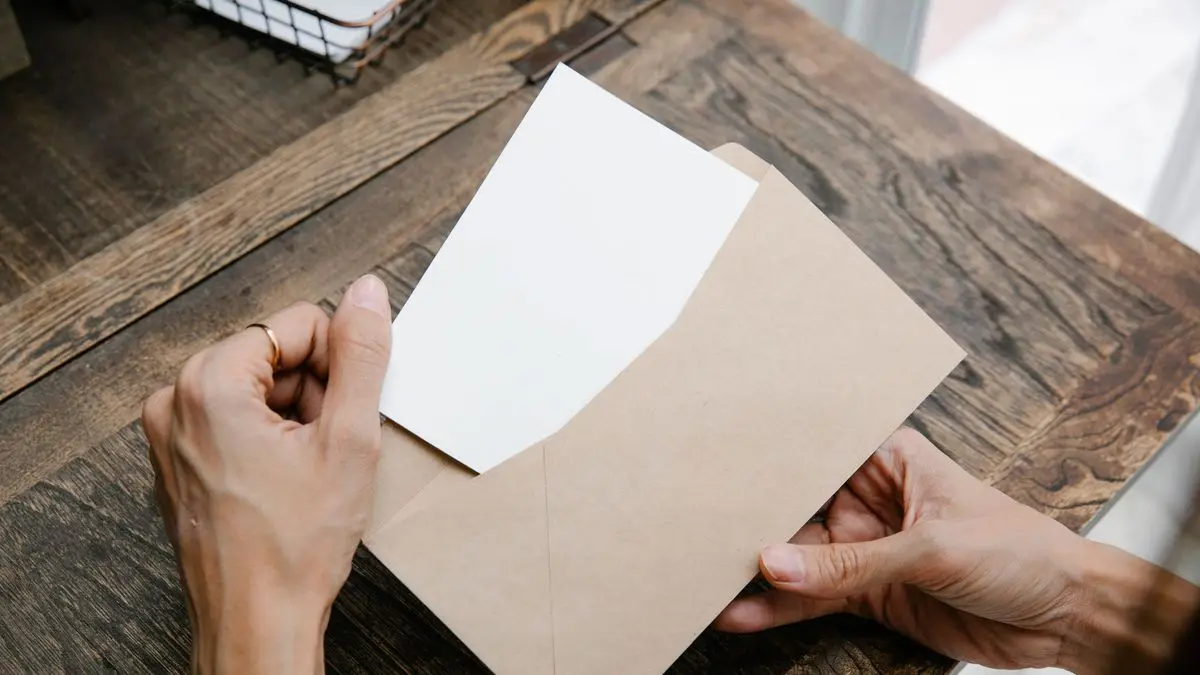 Zwei Hände ziehen auf einem Holztisch ein Blatt Papier aus einem braunen Briefumschlag